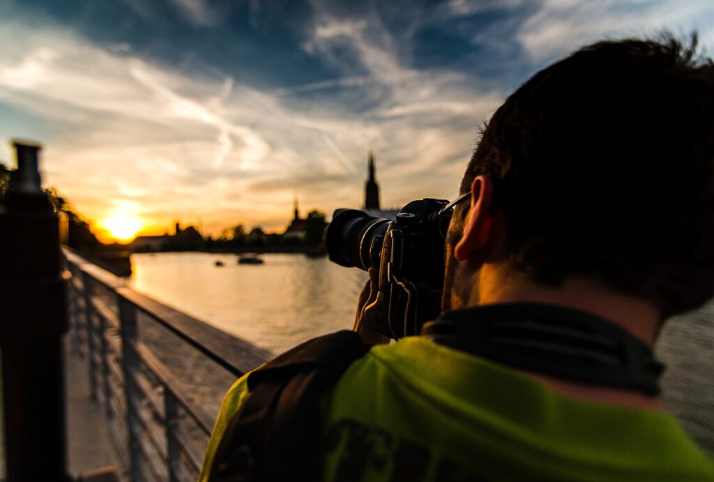 A man in a green t-shirt taking a photo of the sunset with his black DSLR camera